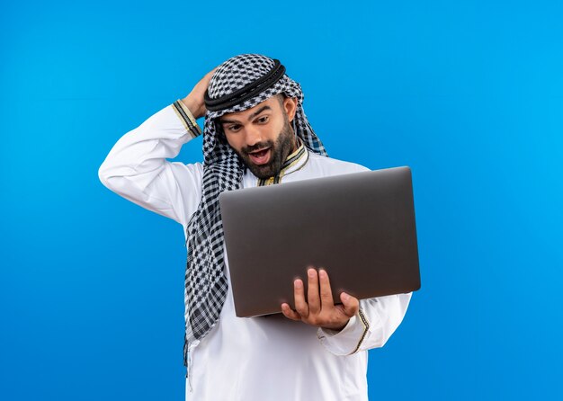 Arabic businessman in traditional wear holding laptop looking at it surprised and happy standing over blue wall