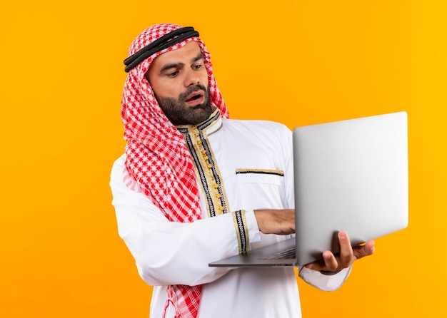 オレンジ色の壁の上に立って驚いて驚いたように見えるラップトップを保持している伝統的な服を着たアラビアのビジネスマン