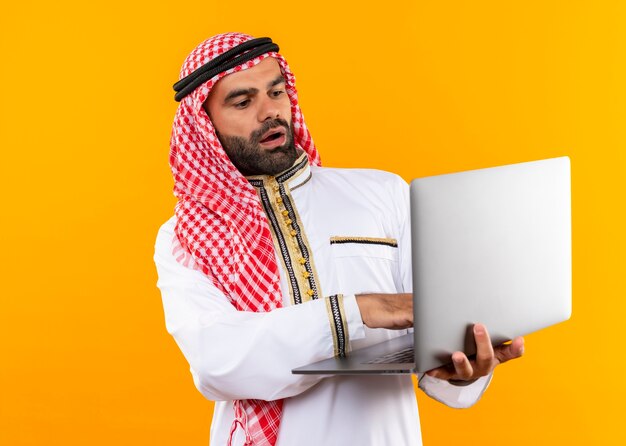 Arabic businessman in traditional wear holding laptop looking surprised and amazed standing over orange wall