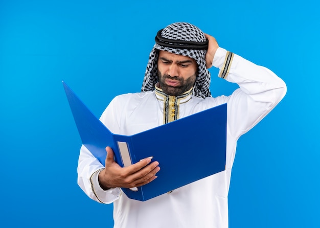 파란색 벽 위에 서있는 머리에 손으로 혼동 그것을보고 오픈 폴더를 들고 전통적인 착용 아랍어 사업가