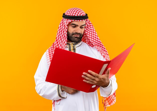 オレンジ色の壁の上に立っている真剣な表情でotを見て開いているフォルダを保持している伝統的な服装のアラビアのビジネスマン