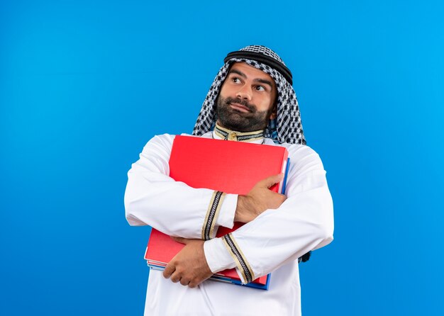 青い壁の上に立って混乱して脇を見て2つのフォルダーを保持している伝統的な服を着たアラビアのビジネスマン