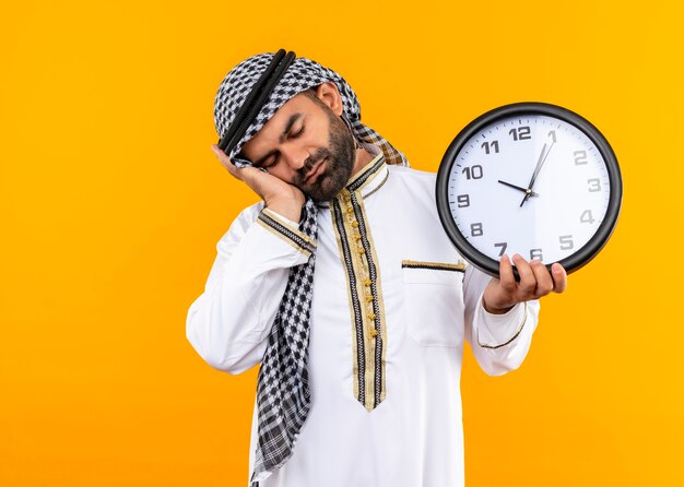 피곤해 보이는 그의 손바닥에 머리를 기대고 벽 시계를 들고 전통적인 착용에서 아랍어 사업가 오렌지 벽 위에 서 자고 싶어