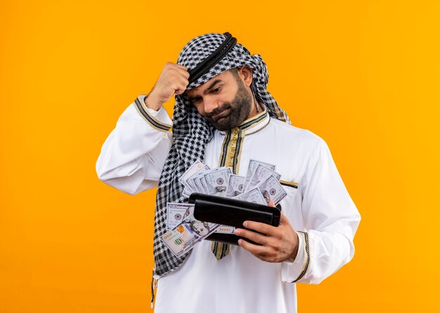 arabic businessman in traditional wear holding wallet with cash looking confused and very anxious standing over orange wall