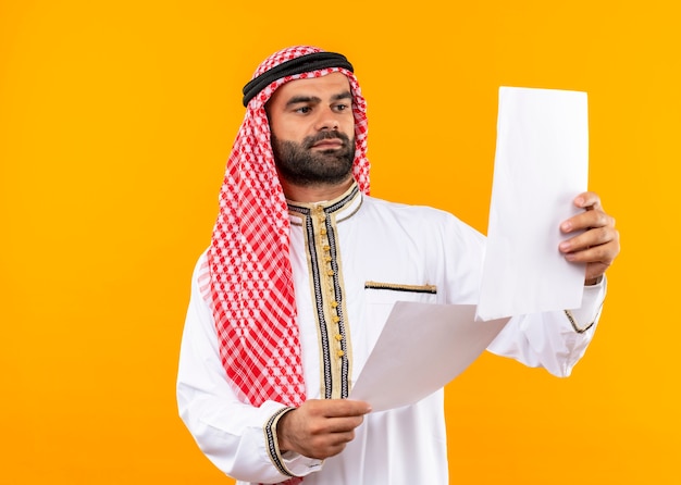 Arabic businessman in traditional wear looking at documentsin hands with serious face standing over orange wall