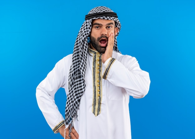 Arabic businessman in traditional wear  shocked touching with arm his face standing over blue wall