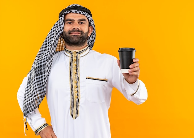 Arabic businessman in traditional wear showing coffee cup with confident smile on face standing over orange wall