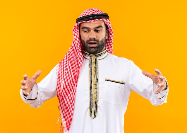 Arabic businessman in traditional wear showing large size gesture with hands, measure symbol, looking surprised standing over orange wall