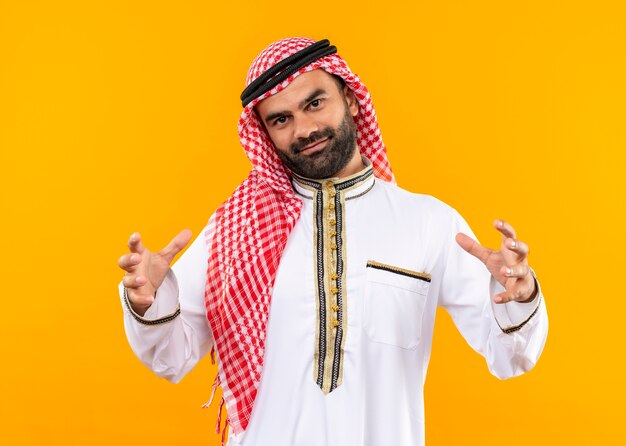 오렌지 벽 위에 서있는 자신감, 측정 기호 미소 손으로 대형 제스처를 보여주는 전통적인 착용 아랍어 사업가