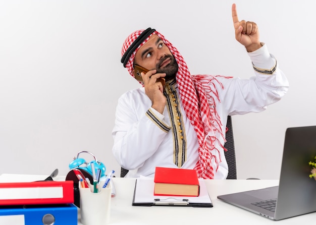 Арабский бизнесмен в традиционной одежде сидит за столом, разговаривает по мобильному телефону, указывая пальцем, работая в офисе