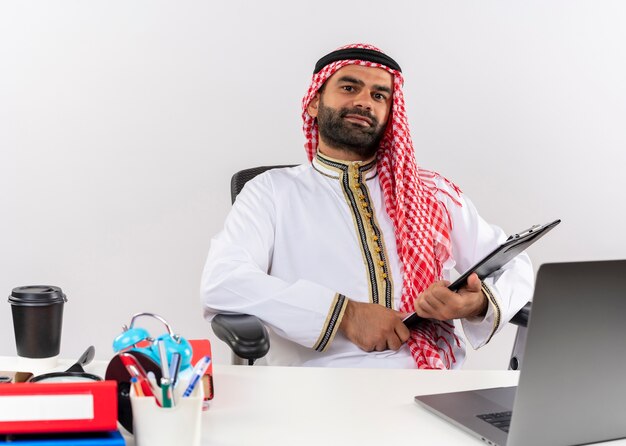Arabic businessman in traditional wear sitting at the table with laptop computer holding clipboard looking confident working in office