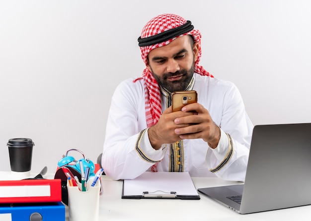 Uomo d'affari arabo nell'usura tradizionale che si siede al tavolo con il computer portatile che tiene il messaggio di sms di smartphone che lavora nell'ufficio