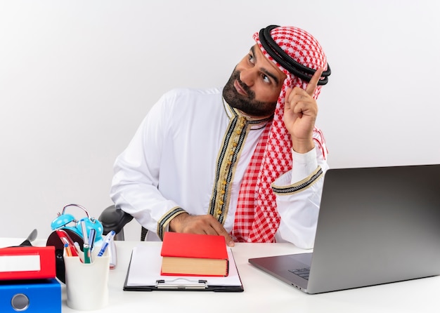 ラップトップコンピューターがオフィスで働いている指を上に向けて脇を向いてテーブルに座っている伝統的な服を着たアラビアのビジネスマン