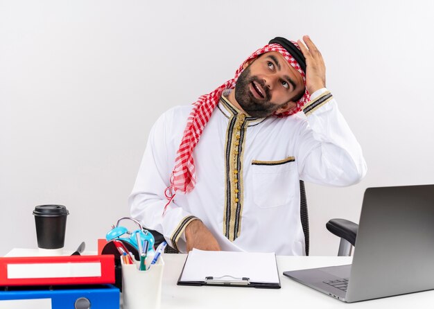 Арабский бизнесмен в традиционной одежде сидит за столом с портативным компьютером, смущенный и сомневающийся в работе в офисе