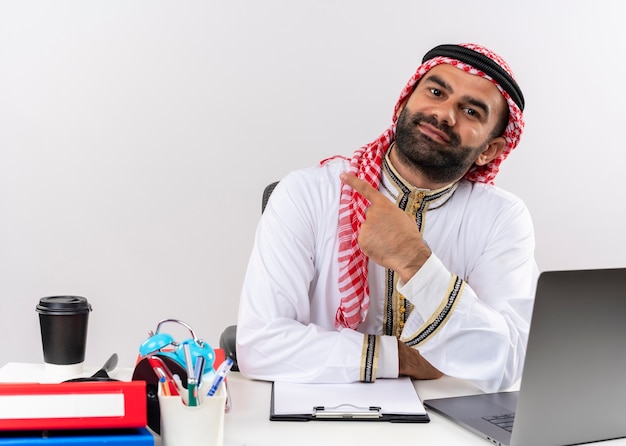 左に人差し指で指しているラップトップコンピューターでテーブルに座っている伝統的な服を着たアラビアのビジネスマンは、オフィスで自信を持って働いているように見えます
