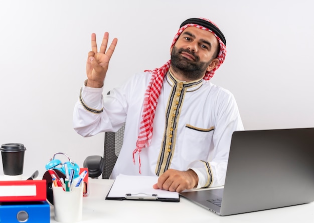 Arabic businessman in traditional wear sitting at the table with laptop computer smiling showing and pointing up with fingers number three working in office