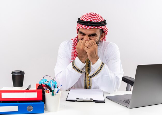 Арабский бизнесмен в традиционной одежде, сидя за столом с портативным компьютером, напряженно и нервно кусает ногти, работая в офисе