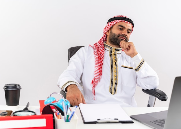 Foto gratuita uomo d'affari arabo in abbigliamento tradizionale seduto al tavolo con computer portatile pensando con espressione pensierosa lavorando in ufficio