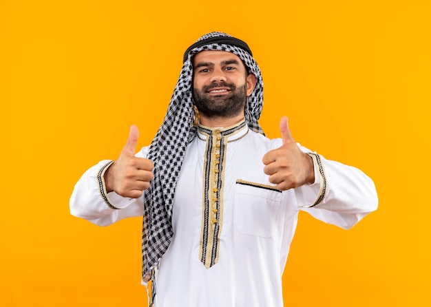 Free photo arabic businessman in traditional wear smiling confident showing thumbs up standing over orange wall