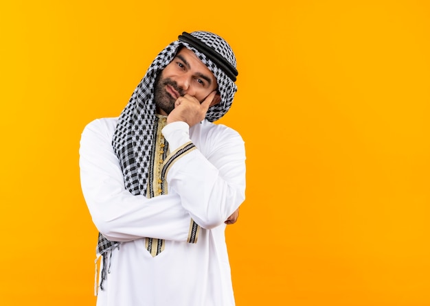 Uomo d'affari arabo nell'usura tradizionale sorridente che sembra stanco e annoiato in piedi sopra la parete arancione