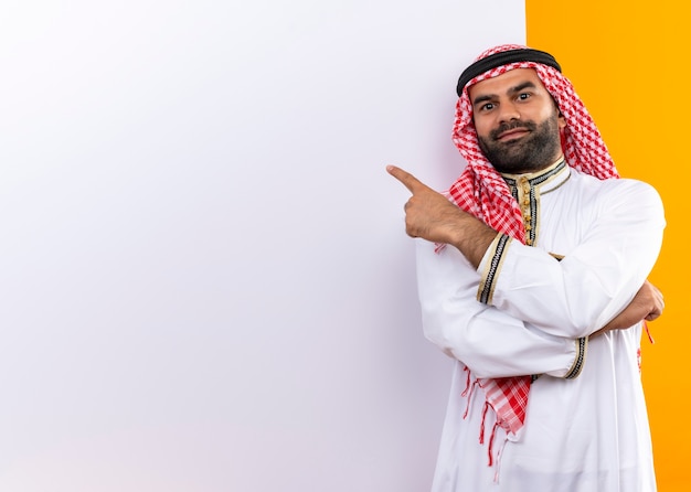 Arabic businessman in traditional wear standing near blank billboard pointing with finger at it with confident smile on face over orange wall