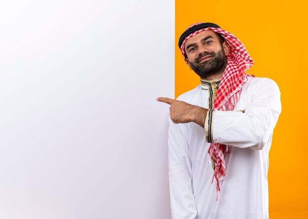 Arabic businessman in traditional wear standing near blank billboard pointing with finger at it with confident smile on face over orange wall