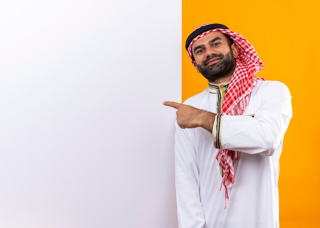 オレンジ色の壁の上の顔に自信を持って笑顔でそれを指で指している空白の看板の近くに立っている伝統的な服を着たアラビアのビジネスマン