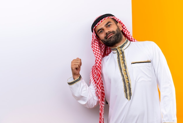 Arabic businessman in traditional wear standing near blank billboard pointing with finger at it with confident smile on face over orange wall