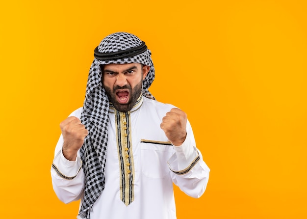 オレンジ色の壁の上に立っている怒っている顔を握りこぶしで伝統的な服装のアラビアの実業家