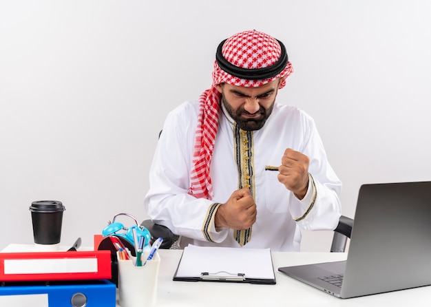 攻撃的な表情で拳を握りしめ、オフィスのテーブルに座って不満と欲求不満のラップトップコンピューターで作業する伝統的な服装のアラビアのビジネスマン