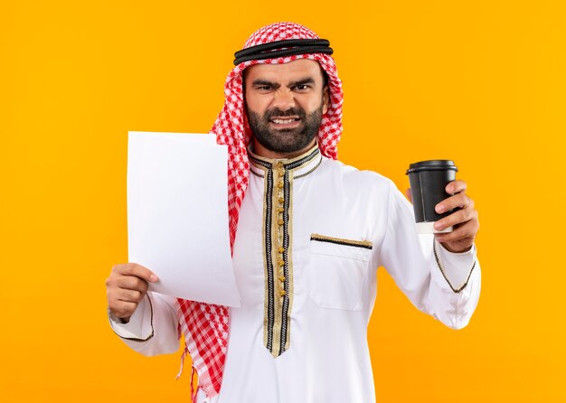 Foto gratuita uomo d'affari arabo nei tradizionali documenti wearholding e tazza di caffè con la faccia arrabbiata in piedi sopra la parete arancione