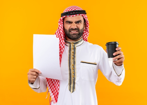 オレンジ色の壁の上に立っている怒っている顔と伝統的なウェアホールディングドキュメントとコーヒーカップのアラビアのビジネスマン