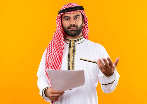 Arabic businessman in traditional wearholding documents  with arm out asking a question standing over orange wall