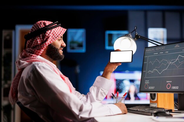 an arabic individual wearing traditional clothing uses a mobile phone at an office desk the isolated white screen offers space for customized content in an advertising or promotional setting
