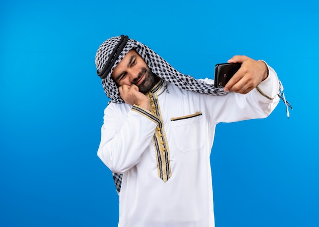 무료 사진 파란색 벽 위에 서있는 행복한 얼굴로 웃는 스마트 폰을 사용하여 셀카를 복용하는 전통적인 착용에 아랍 사람