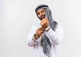 Free photo arabic man in traditional wear fixing his cufflink with confident look standing over white wall