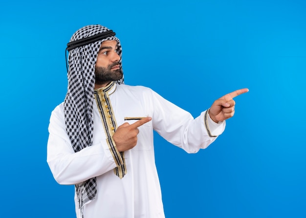 Arabic man in traditional wear looking aside pointing with fingers to the side with serious face standing over blue wall