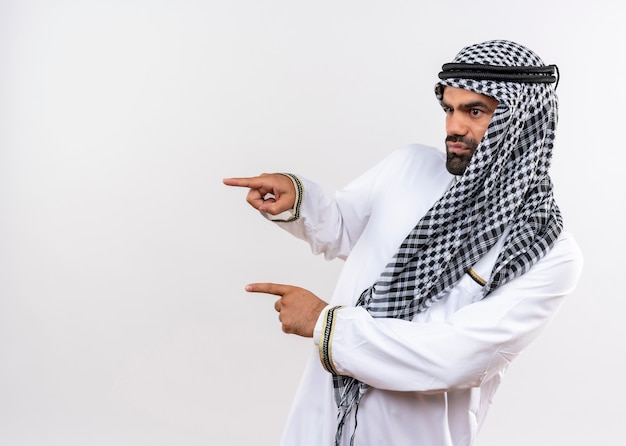Arabic man in traditional wear looking aside with serious face pointing with fingers to the side standing over white wall