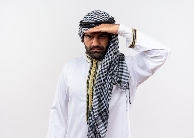 Арабский мужчина в традиционной одежде смотрит вдаль с рукой над головой, стоя над белой стеной