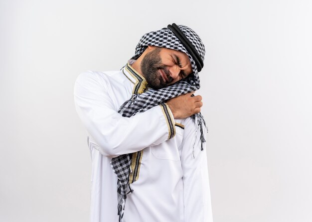 Arabic man in traditional wear looking unwell touching shoulder having pain standing over white wall