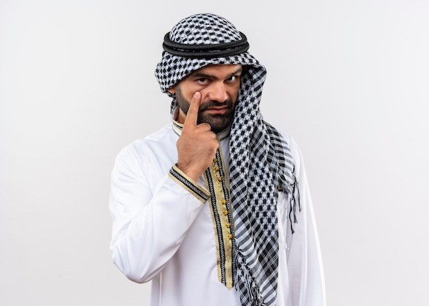 Free photo arabic man in traditional wear pointing to his eye with confident expression standing over white wall