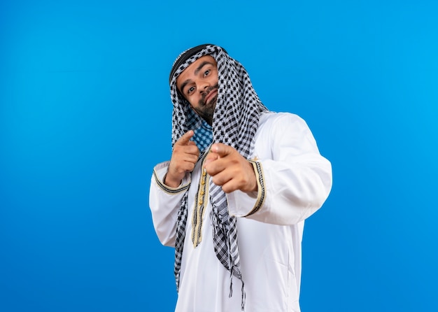 파란색 벽 위에 서있는 얼굴에 자신감이있는 미소로 손가락으로 가리키는 전통적인 착용에 아랍 사람