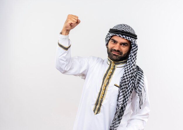 Арабский мужчина в традиционной одежде, поднимающий кулак, уверенно улыбаясь, стоит над белой стеной