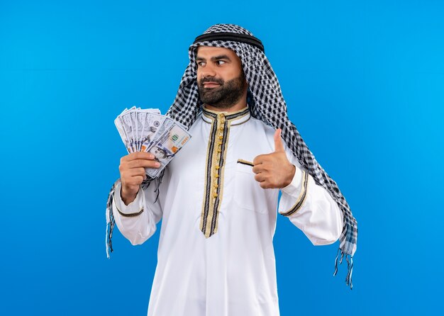 arabic man in traditional wear showing cash looking aside smiling confident showing thumbs up standing over blue wall