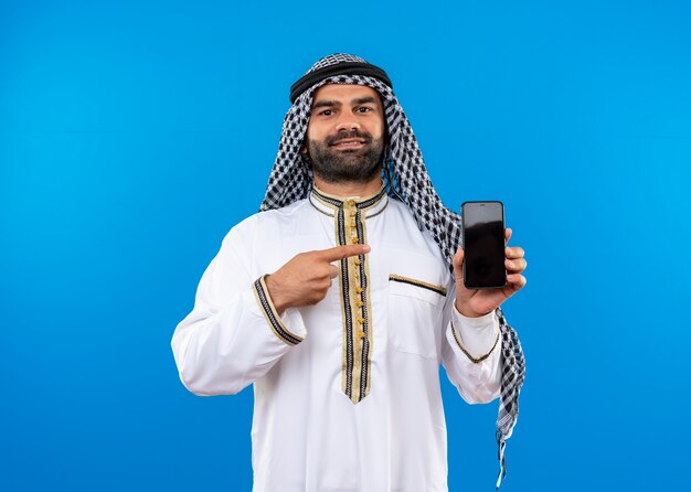 Арабский мужчина в традиционной одежде показывает смартфон, указывая пальцем на него, уверенно улыбаясь, стоя над синей стеной