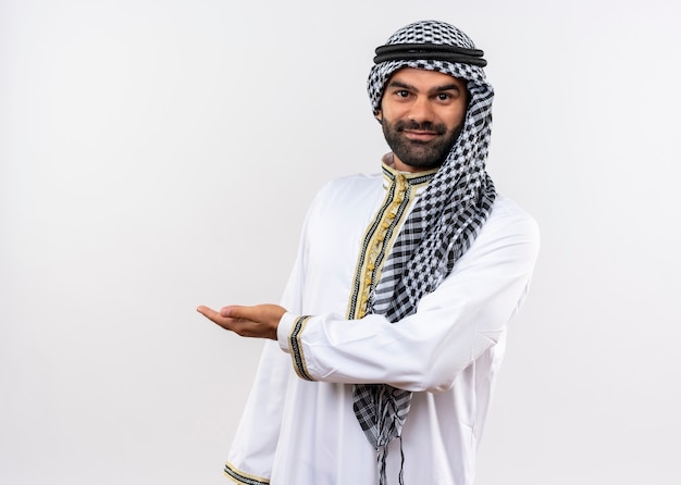 Uomo arabo in abbigliamento tradizionale sorridente prsenting con il braccio della mano in piedi sul muro bianco