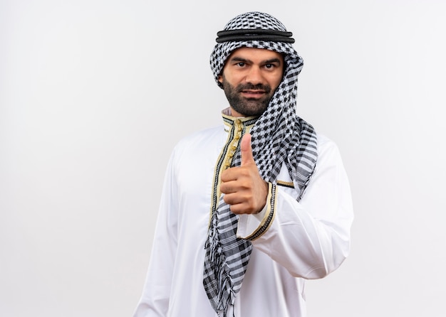 Арабский мужчина в традиционной одежде улыбается, показывает палец вверх, стоя над белой стеной