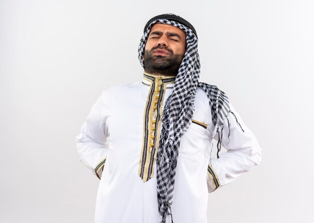 Arabic man in traditional wear touching back looking unwell having pain standing over white wall