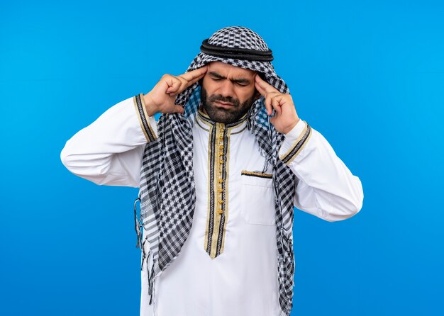 Arabic man in traditional wear touching his temples suffering from strong headache standing over blue wall