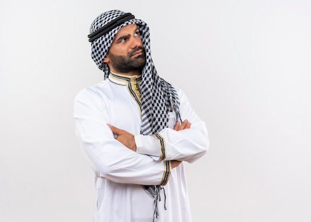 Free photo arabic man in traditional wear with crossed arms on chest looking aside with serious face standing over white wall